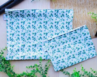 Pode incluir: Duas sacolas de compras reutilizáveis com fundo branco e um padrão de folhas de eucalipto verdes. As sacolas são feitas de um tecido macio e durável e são perfeitas para carregar mantimentos ou outros itens.