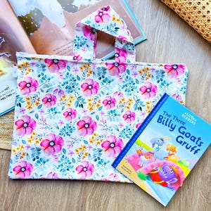 Op de afbeelding: Een witte boekentas met een bloemenprint met roze bloemen en groene bladeren. De tas heeft een handvat en bevat een kinderboek met de titel "The Three Billy Goats Gruff" met een blauwe omslag en kleurrijke illustraties.