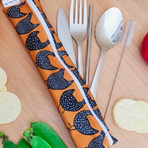 May include: An orange and white chicken print cutlery case with a white zipper, containing a knife, fork, spoon, chopsticks, and a straw cleaner. Fresh green pea pods, orange, and potato chips are arranged around the case.