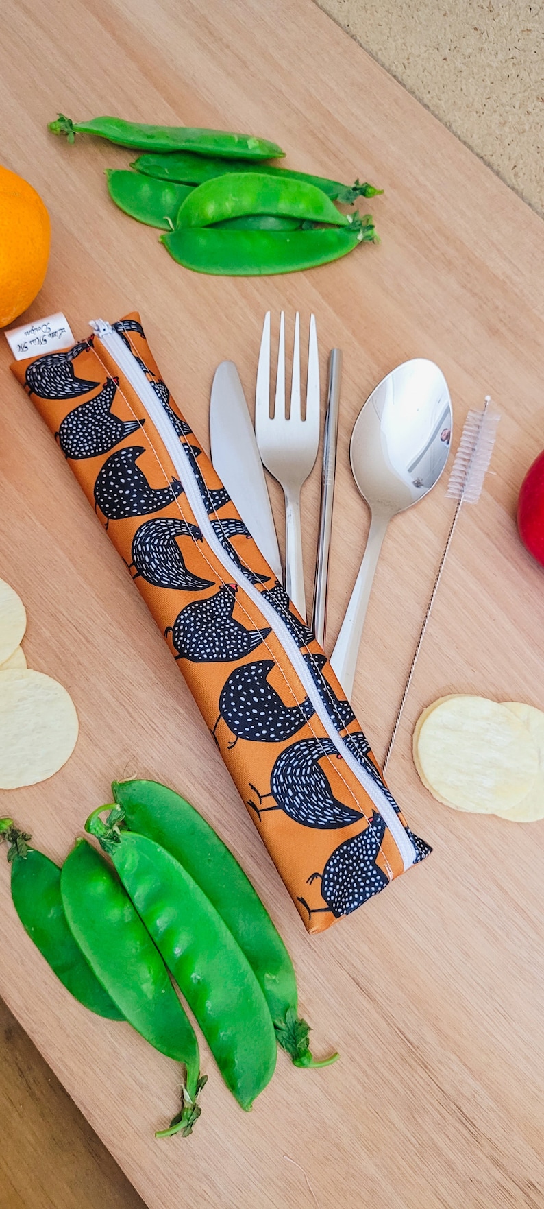 May include: An orange and white chicken print cutlery case with a white zipper, containing a knife, fork, spoon, chopsticks, and a straw cleaner. Fresh green pea pods, orange, and potato chips are arranged around the case.