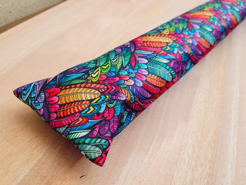 May include: A colorful, long, rectangular pillow with a pattern of feathers in shades of red, orange, yellow, green, blue, purple, and pink on a black background.