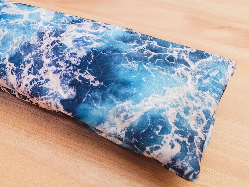 May include: A blue and white patterned eye pillow with a wave design. The pillow is made of soft fabric and is filled with flax seeds and lavender.