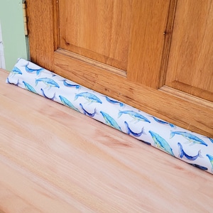 May include: A white doorstop with a blue whale pattern. The doorstop is made of fabric and is filled with beans. It is placed at the bottom of a wooden door.