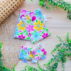 May include: Three colorful fabric bibs with a floral pattern. The bibs are in different sizes and feature a white background with pink, yellow, blue, and green flowers. The bibs are made by "Little Miss Designs".
