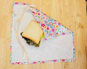 Puede incluir: Un envoltorio de sándwich de tela blanca con estampado floral en tonos rosa, morado y azul. El envoltorio está doblado sobre un sándwich con lechuga y pan blanco.