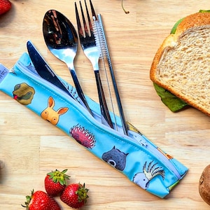 Könnte beinhalten: Eine blaue Reißverschlusstasche mit einem Cartoon-Tierdruckdesign. Die Tasche enthält eine Gabel, ein Messer, einen Löffel und eine Reinigungsbürste. Die Tasche befindet sich auf einer Holzoberfläche mit einem Sandwich, Obst und anderen Gegenständen.