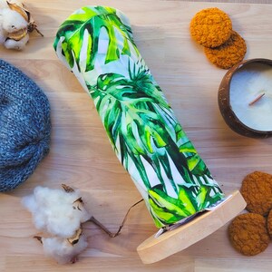 May include: A white fabric tube with a green tropical leaf print. The tube is rolled up and tied with a wooden dowel. The tube is sitting on a wooden surface.