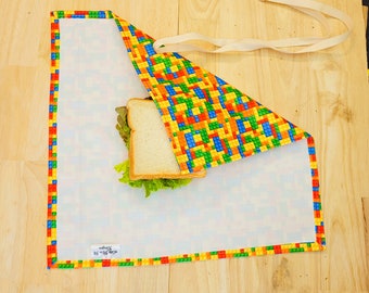 Puede incluir: Un envoltorio reutilizable para sándwiches colorido con un patrón tipo Lego. El envoltorio está doblado sobre un sándwich y tiene una etiqueta blanca que dice "sandwich wrap 12" x 14" ".