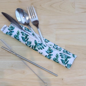 May include: A white zippered pouch with a green leafy pattern holds a silver fork, knife, and spoon. A silver drinking straw with a cleaning brush lies on a light wood surface.