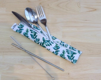 May include: A white zippered pouch with a green leafy pattern holds a silver fork, knife, and spoon. A silver drinking straw with a cleaning brush lies on a light wood surface.