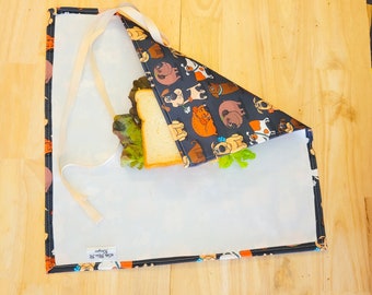 May include: A reusable sandwich wrap featuring a black background with various dog illustrations in brown, orange, and white. The wrap is open, revealing a sandwich with lettuce. The wrap has a white interior and beige ties.