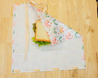 Puede incluir: Un envoltorio de comida reutilizable blanco con un patrón de arcoíris y estrellas. El envoltorio está doblado sobre un sándwich con lechuga y pan blanco.