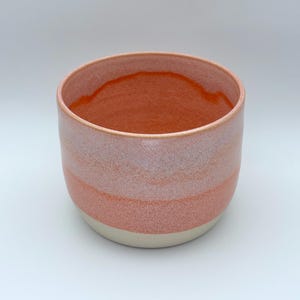 May include: A ceramic bowl with a gradient of colours. The interior is a deep orange, transitioning to a lighter orange and pink on the exterior. The base is a neutral off-white colour.