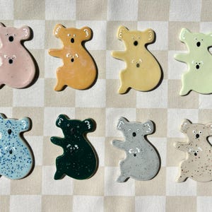 May include: Eight ceramic koala figurines in various colours, including pink, yellow, light green, blue, green, grey, and beige. Each koala is sitting with its arms outstretched and has a small, white koala inside its belly.