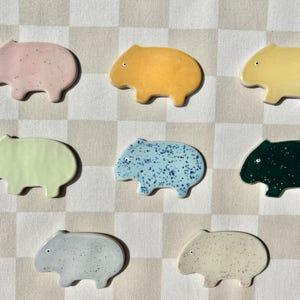May include: Set of ten ceramic animal figurines in various colours, including pink, yellow, green, blue, and white. Each figurine is shaped like a wombat and has a unique pattern.