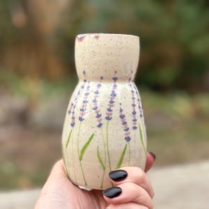 May include: A speckled, cream-coloured ceramic vase with a lavender floral design. The vase has a wide top and a narrower base, with purple and green painted details. The vase is held in a hand.