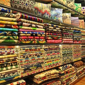 Op de afbeelding: Meerdere planken gestapeld met gevouwen textiel en decoratieve kussens met diverse ikat-patronen in levendige kleuren, waaronder blauw, rood, groen, geel en zwart. De stoffen zijn netjes in rijen gerangschikt en tonen diverse geometrische en abstracte ontwerpen.