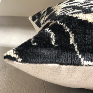 Op de afbeelding: Een vierkant sierkussen met een contrastrijk zwart-wit abstract patroon op de voorzijde. De stof heeft een getextureerde, fluweelachtige pool, terwijl de zijnaad en het achterpaneel zijn gemaakt van een effen, lichtbeige geweven textiel.