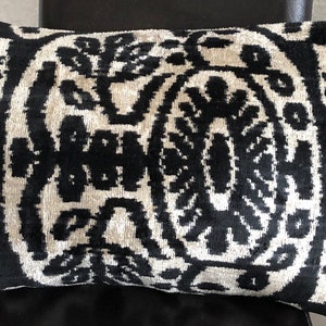 Op de afbeelding: Een rechthoekig sierkussen met een contrastrijk ikat-patroon in zwart en gebroken wit. Het ontwerp bestaat uit complexe, symmetrische en abstracte bloemmotieven die zich herhalen over het gehele oppervlak van de fluweelachtige stof.