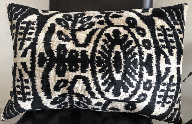 Op de afbeelding: Een rechthoekig sierkussen met een contrastrijk ikat-patroon in zwart en gebroken wit. Het ontwerp bestaat uit complexe, symmetrische en abstracte bloemmotieven die zich herhalen over het gehele oppervlak van de fluweelachtige stof.