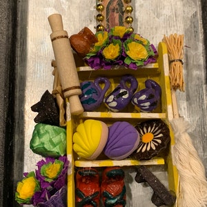 Può includere: Uno scaffale di legno giallo con vari oggetti di artigianato popolare messicano, tra cui un mattarello, un vaso di terracotta, un'icona religiosa, fiori e una scopa. Lo scaffale è decorato con vernice bianca e ha uno sfondo nero.