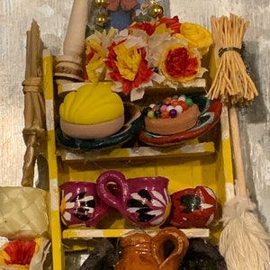 Può includere: Uno scaffale di legno giallo con vari oggetti decorativi, tra cui una piccola scopa, un vaso di terracotta, una torta gialla, una piccola cornice con un'immagine religiosa e un bouquet di fiori gialli e rossi.