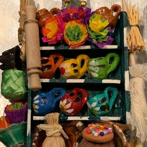 Può includere: Un ripiano in legno in miniatura con tre ripiani, decorato con fiori colorati, vasi di terracotta e altri oggetti artigianali messicani tradizionali. Il ripiano è dipinto di verde e bianco.