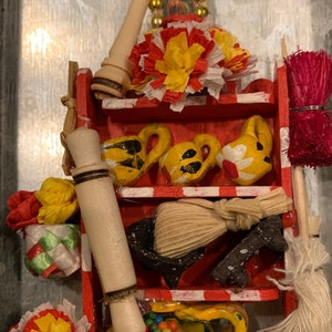 Può includere: Uno scaffale in legno rosso con vari oggetti, tra cui un mattarello, una scopa, un piccolo cestino con perline colorate e un vaso in ceramica giallo con un manico marrone. Lo scaffale è decorato con strisce bianche e rosse e ha un fiore giallo in cima.