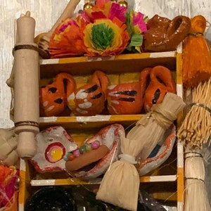 Può includere: Un ripiano in legno in miniatura con vari oggetti di artigianato popolare messicano, tra cui un mattarello in terracotta, un vaso in terracotta, una ciotola in terracotta, una figurina in terracotta e una piccola immagine incorniciata di una figura religiosa. Il ripiano è decorato con fiori di carta colorati e una collana di perline dorate.