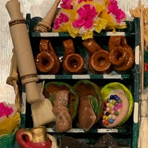 Può includere: Un ripiano in legno in miniatura con un design a strisce verde e bianco. Il ripiano è decorato con una varietà di oggetti, tra cui un mattarello, un piccolo pennello di legno, una tazza di ceramica, un piccolo brocca di ceramica e una piccola ciotola di ceramica. Il ripiano è anche decorato con un piccolo bouquet di fiori rosa e gialli, una piccola immagine di una figura religiosa e una piccola collana di perle.