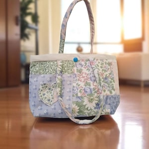 May include: A patchwork tote bag with floral patterns in shades of blue, green, and pink. The bag features a heart-shaped appliqué and a blue button closure. The handles and trim are a complementary light pink.
