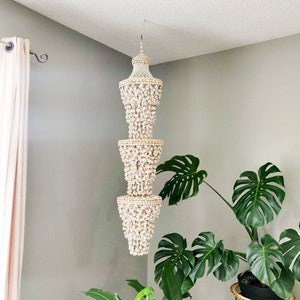 May include: A three-tiered chandelier made of seashells hangs from the ceiling. The chandelier has a cream-colored hue and is suspended in a corner of a room with gray walls. Green plants are in the foreground.