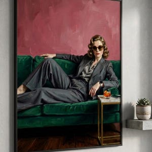May include: A framed art print featuring a person in a gray suit lounging on a green velvet sofa. The background is a textured red-pink color. A small side table holds a drink and a phone. The artwork is a stylish, modern portrait.