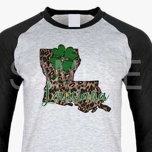 May include: A gray and black baseball style t-shirt with a leopard print outline of the state of Louisiana with a green four-leaf clover on top. The text "Louisiana" is written in black cursive below the state outline. The text "DIGITAL DESIGN" is in a white oval with a black border.