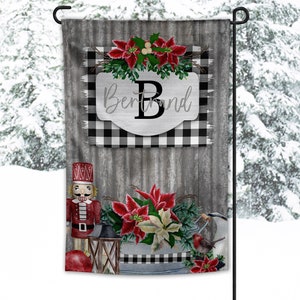May include: A decorative garden flag with a winter theme. The flag features a black and white buffalo check pattern, poinsettias, and a nutcracker. The word "Brand" is displayed with the letter "B". The background is a corrugated metal design.
