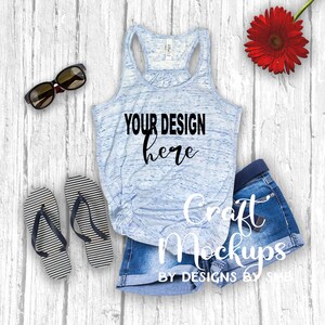 May include: A light blue tank top with the text "YOUR DESIGN here" is displayed with denim shorts, sunglasses, and striped flip-flops. A red gerbera daisy adds a pop of color. The items are arranged on a white wood-grain surface.