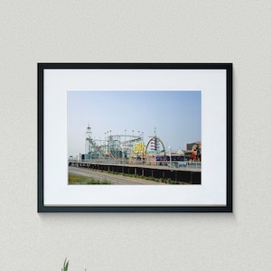 Op de afbeelding: Een ingelijste foto van een kleurrijk pretpark met een achtbaan en andere attracties tegen een helderblauwe lucht. De zwart omlijste foto hangt aan een witte muur, boven een cactus in een pot en een zwarte Marshall-luidspreker.