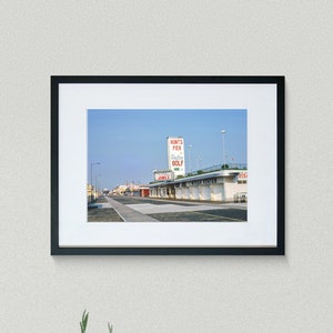 May include: A black and white photograph of a building with a sign that reads "Runt's Pier, Bathing, Golf". The building is situated on a street with a few cars parked in front of it.