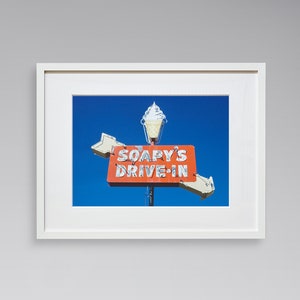 May include: A white framed photograph of a vintage neon sign with a blue sky background. The sign reads "Soapy's Drive-In" and features a stylized ice cream cone.