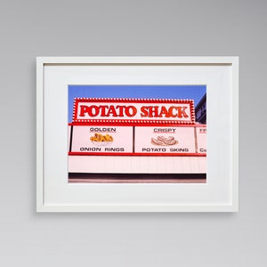 May include: A white framed photograph of a red and white sign that reads "Potato Shack" with a blue sky in the background. The sign advertises "Golden Onion Rings" and "Crispy Potato Skins".