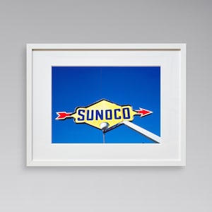 May include: Framed print featuring a vintage Sunoco sign against a bright blue sky. The sign is yellow with red arrows and the word "SUNOCO" in bold, blue letters. The print is in a white frame.