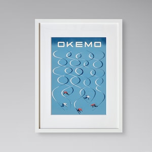 May include: A framed vintage poster with the word "OKEMO" at the top. The poster features skiers on a blue background with white looping lines. The frame is white, and a large green leaf is in the foreground.