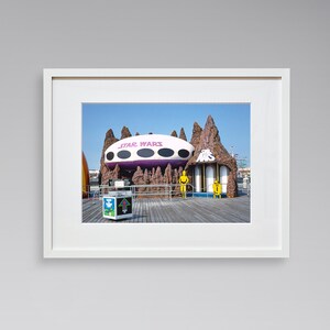 May include: A framed photograph of a Star Wars themed amusement park ride. The ride is a large, white, flying saucer with purple accents. The saucer is hovering over a brown, rocky landscape. The words "Star Wars" are printed in pink on the saucer.