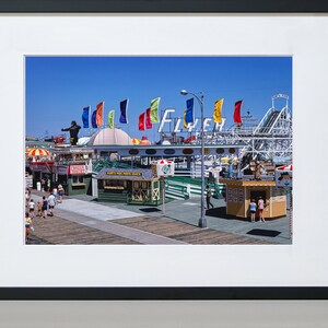 Puede incluir: Una fotograf&iacute;a vintage de un parque de atracciones en un paseo mar&iacute;timo con una monta&ntilde;a rusa, banderas de colores y un letrero que dice "Flyer". Hay gente caminando por el paseo mar&iacute;timo y varios puestos de comida.