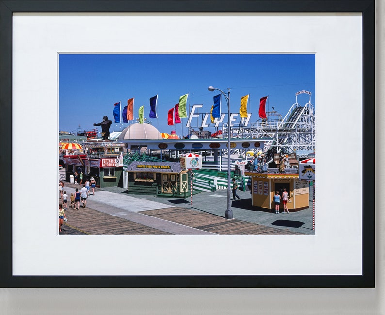 Puede incluir: Una fotograf&iacute;a vintage de un parque de atracciones en un paseo mar&iacute;timo con una monta&ntilde;a rusa, banderas de colores y un letrero que dice "Flyer". Hay gente caminando por el paseo mar&iacute;timo y varios puestos de comida.