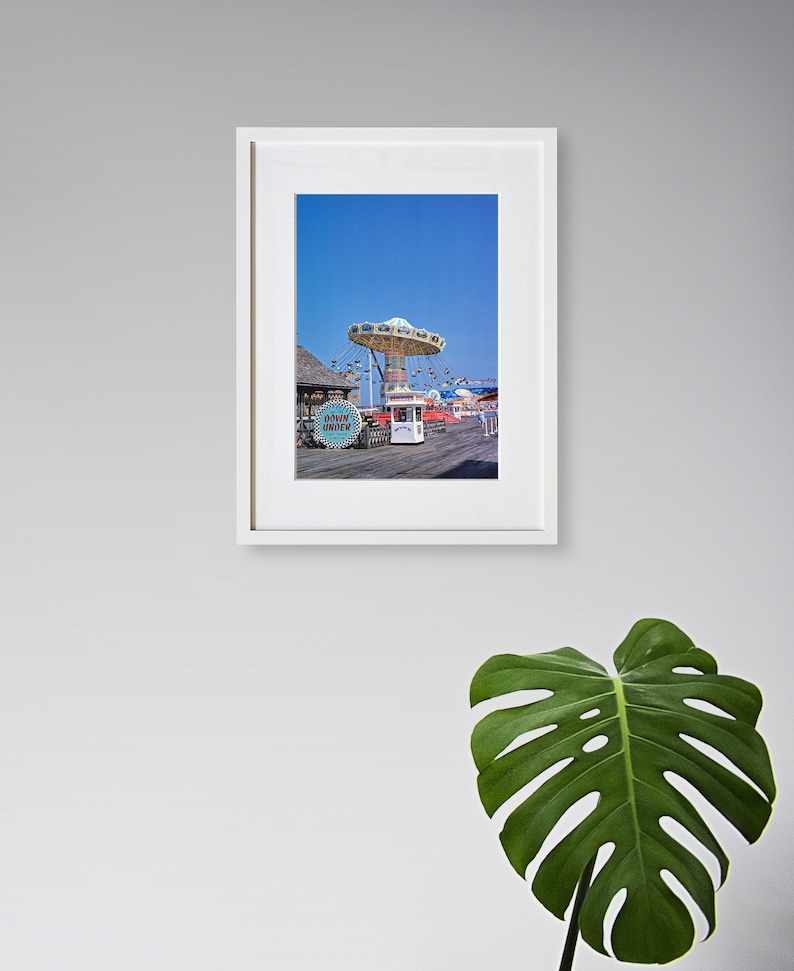 May include: A framed photograph of a carousel ride on a boardwalk with a blue sky in the background. The carousel is white with red and blue accents. The sign "Down Under" is visible in the foreground.