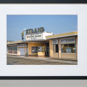 K&ouml;nnte beinhalten: Ein Vintage-Foto eines Kinos namens "The Strand" mit einem Leuchtkasten, der "Sylvester Stallone Fist" bewirbt, das um 19:25 Uhr und 22:10 Uhr spielt. Nebenan befindet sich ein Restaurant namens "Spire's Restaurant".