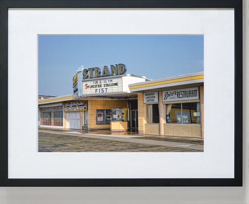 K&ouml;nnte beinhalten: Ein Vintage-Foto eines Kinos namens "The Strand" mit einem Leuchtkasten, der "Sylvester Stallone Fist" bewirbt, das um 19:25 Uhr und 22:10 Uhr spielt. Nebenan befindet sich ein Restaurant namens "Spire's Restaurant".