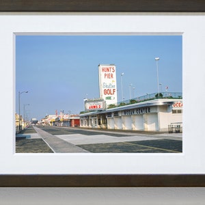 May include: A vintage photograph of a street scene in a seaside town. The photo shows a street lined with buildings, including a building with a sign that reads "Hunt's Pier Skyline Golf". The street is empty and the sky is blue.