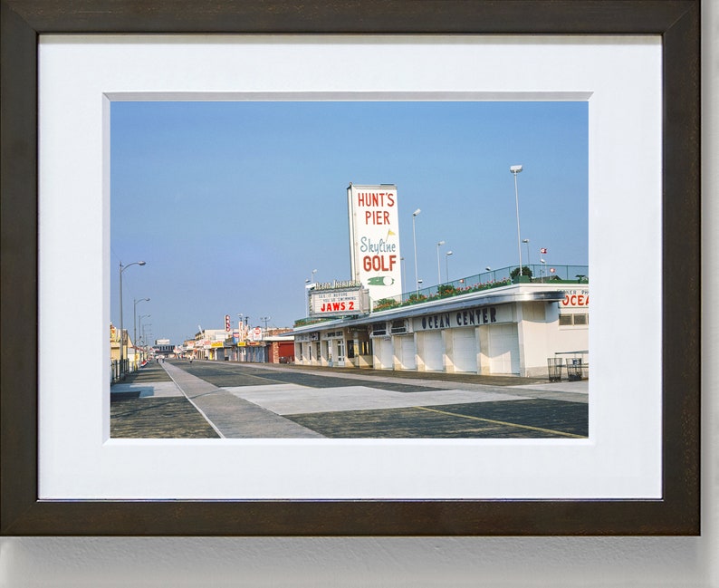 May include: A vintage photograph of a street scene in a seaside town. The photo shows a street lined with buildings, including a building with a sign that reads "Hunt's Pier Skyline Golf". The street is empty and the sky is blue.